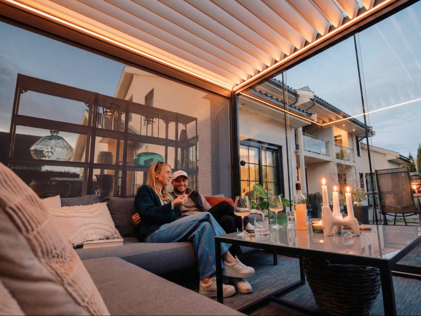 A completed PergoLux Pergola 4 Pro installation at dusk: motorized louvered roof with beam lighting, glass side walls and outdoor sofa on a finished terrace